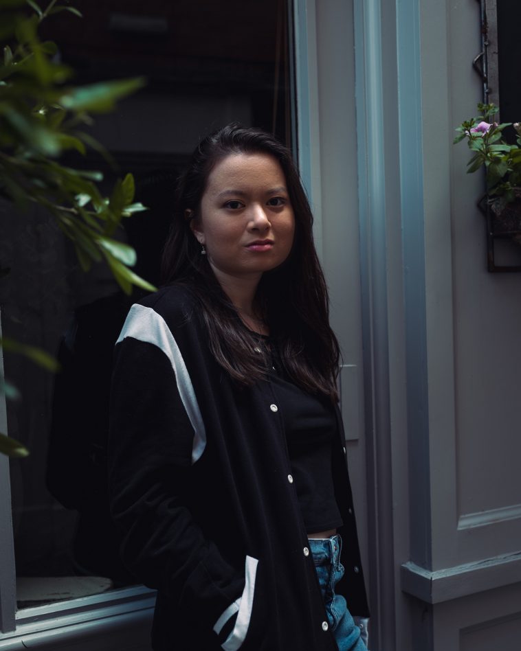 Promotional photo for "Running" which sees Laura Loh leaning on a monotone blue windowsill, against a window which has a tall plant behind. She is wearing a black varsity jacket that has a white stripe at the top of the sleeve and at the bottom, over a black t-shirt and blue jeans with her long dark-brown hair worn down past her shoulders.