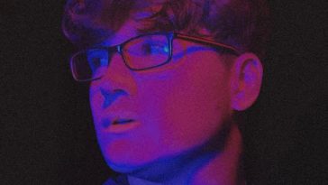 Promotional photo for "Far" which sees a head-shot of Rob Earle as he's looking to his right, with light reflecting off his glasses. He has short brown hair with a curly fringe, and he's wearing a grey sweater.