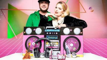 Promotional photo for the Halloween Playlist which sees Charity Daw and Josh Edmondson pose in front of a pink background with a huge boombox on a table in front of them.