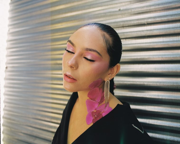 Promotional photo for "the people look like flowers (at last)" which sees Alejandra against a metal garage door, with her hairtail behind her, and she has pink flowers up the left-side of her neck, which is facing us. Her eyes are closed, almost as if she's basking in the sunlight.