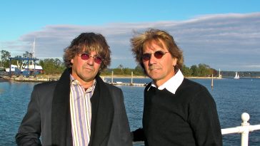 Promotional photo for "Dreams Come True" which see twin-brothers the Alessi Brothers posing against a white railing looking over the sea with a beach in the background and a bright blue sky. They both have bowl-cut-like haircuts and are wearing shades, with the one on the right wearing a white shirt under a black jumper and the brother on the left wearing a white shirt under a navy suit jacket and a black scarf.