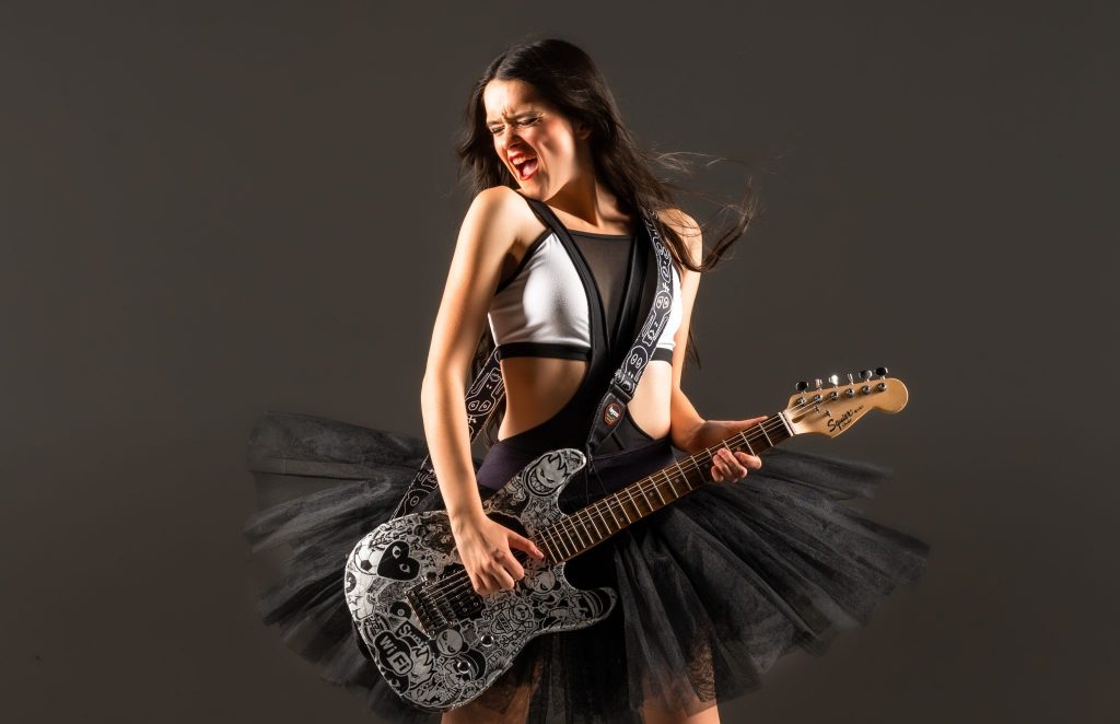 Promotional photo for "Just B U" which sees Belle posing with her glittery black and silver guitar facing head-on with her face turned to the left as a wind machine flows her black hair behind her as well as lifts her black tutu up slightly. A grey background is behind her.