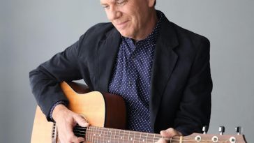 Promotional image for "When Dreams End" which sees Chris St. John sitting on a black bench with a grey wall behind him, playing a guitar. He is looking down at the guitar, wearing a suit jacket, a shirt and a pair of jeans.