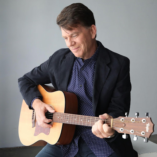 Promotional image for "When Dreams End" which sees Chris St. John sitting on a black bench with a grey wall behind him, playing a guitar. He is looking down at the guitar, wearing a suit jacket, a shirt and a pair of jeans.
