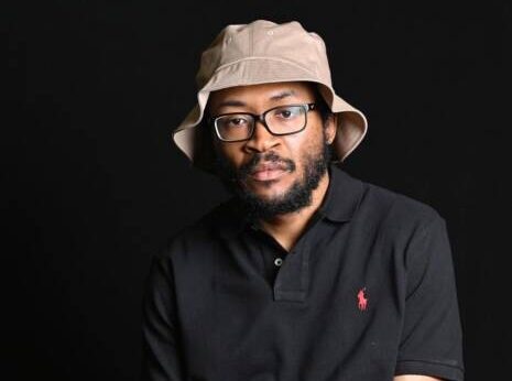 Tea Sea sits in front of a black background, wearing a cream-grey bucket hat, paired with glasses and a black Ralph Lauren polo shirt.