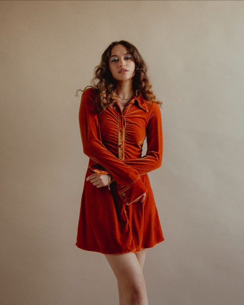 Promotional photo for "Everything You Wish For" which sees Eva Rose posing with her arms crossed in front of her, wearing a dark orange dress, backed by a grey background.