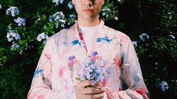 Promotional photo for "ren" which sees Jiubel standing in front of a tall hedge, wearing a pink flower-patterned shirt, holding a bunch of sky blue flowers together in his hands.