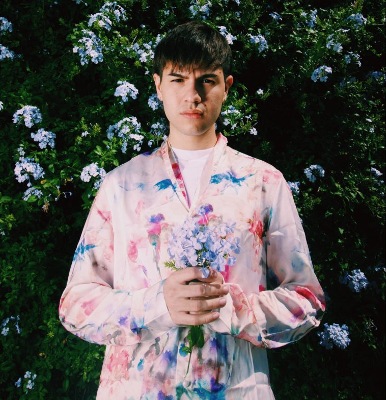 Promotional photo for "ren" which sees Jiubel standing in front of a tall hedge, wearing a pink flower-patterned shirt, holding a bunch of sky blue flowers together in his hands.
