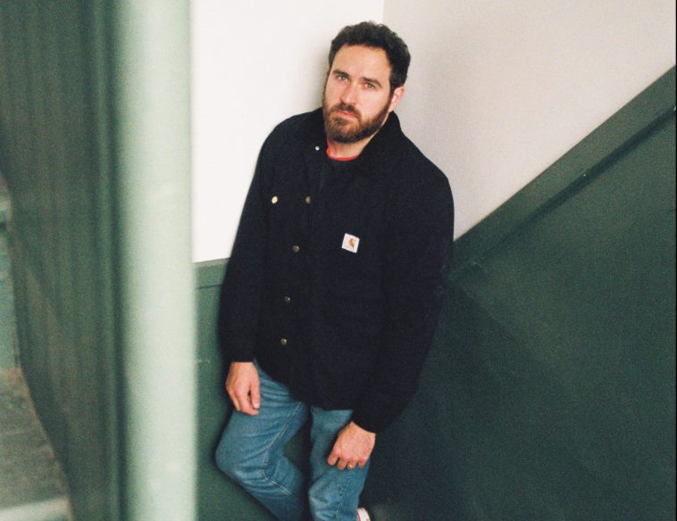 Promotional photo for "When We Were Kids Ourselves" which sees Stephen Babcock leaning against a corner in a stairwell that is sectioned with white walls above and deep green walls below. He is wearing a black hoodie and blue jeans.