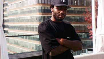 Promotional photo for "Beach Dreams" which sees wallyPDF standing on a rooftop floor of a building overlooking a skyscraper behind him, with his arms crossed over his black t-shirt. He is wearing a navy blue sports cap.
