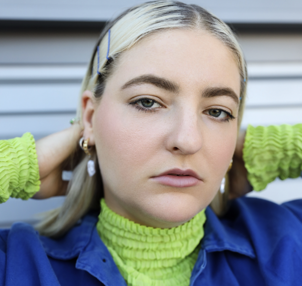 Promotional photo for "TO BE YOUR FRIEND" which sees JORDI UP LATE with slicked back blonde hair that reaches down to her shoulders, paired with metal clips at the side of her temple. Her makeup is perfection and she is wearing a green sweater under a blue shirt-jacket.