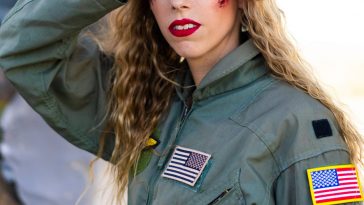 Promotional photo for "Skydive" which sees Juliette Irons wearing dark green pilot overalls with USA flag stitched badges. Her curly dirty-blonde hair fall past her shoulders and she's looking at the camera with her right hand against her forehead in a salute pose. She has wounds on her cheeks and to the side of her right eye.