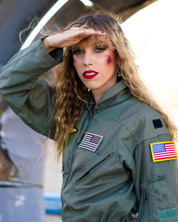 Promotional photo for "Skydive" which sees Juliette Irons wearing dark green pilot overalls with USA flag stitched badges. Her curly dirty-blonde hair fall past her shoulders and she's looking at the camera with her right hand against her forehead in a salute pose. She has wounds on her cheeks and to the side of her right eye.