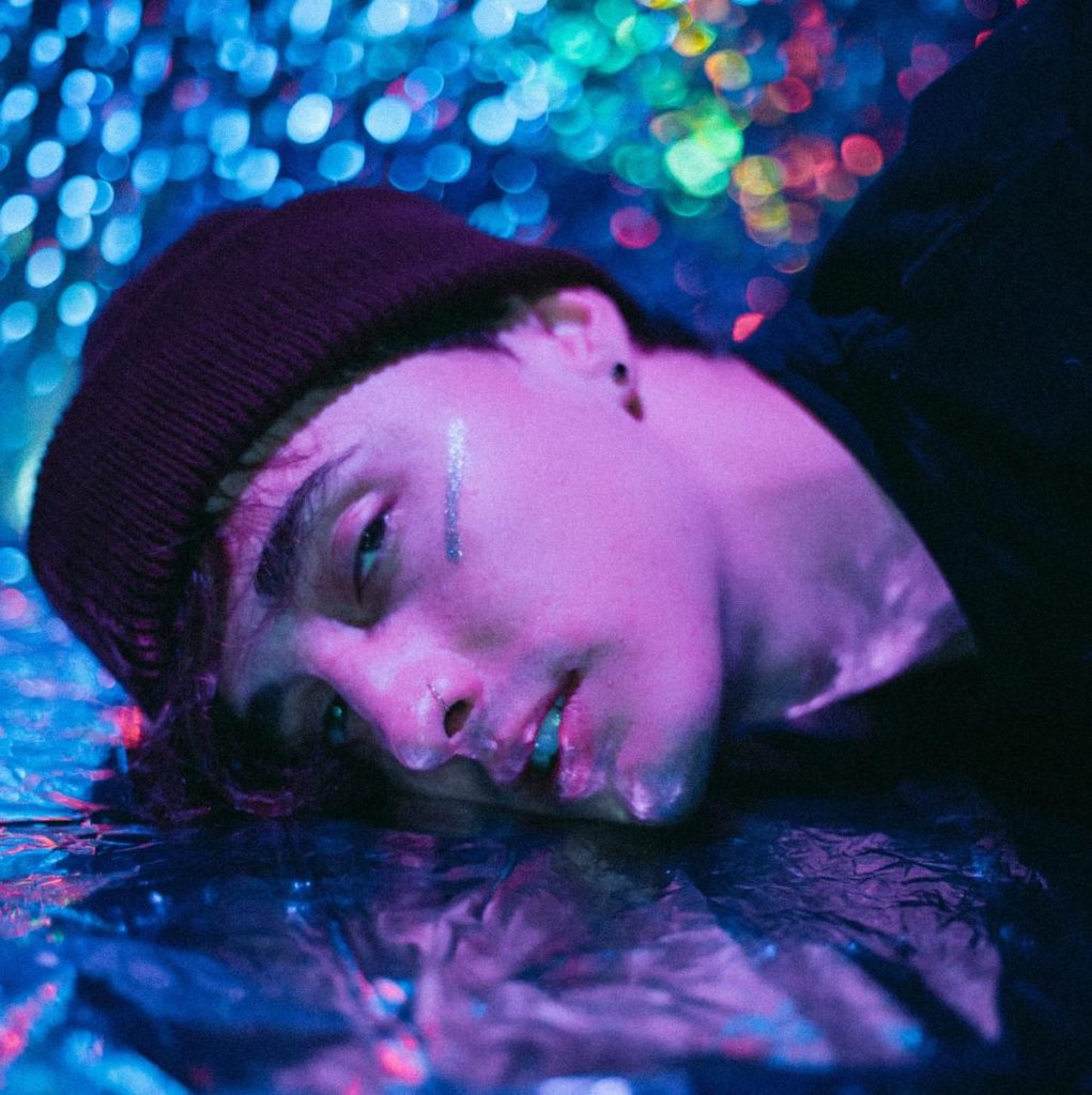 Promotional photo for "Aaron" which sees a headshot of Marcaux lying down on a mirrored metal floor with similar metal walls behind him. He's wearing a beenie hat and a black t-shirt. We can only see his left-side of his face, and there's a silver line under his eye at the top of his cheek. He has neck tattoos and looks defeated.