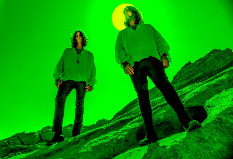 Promotional photo for "Desert Island" which sees Royal Prismatic standing on a sloped rock hill with the sun or moon behind one of the duo. They are both wearing black jeans, boots and pale hoodies. The image has a bright green filter to it, and the male duo's hair are shoulder-length, somewhat covering their faces.