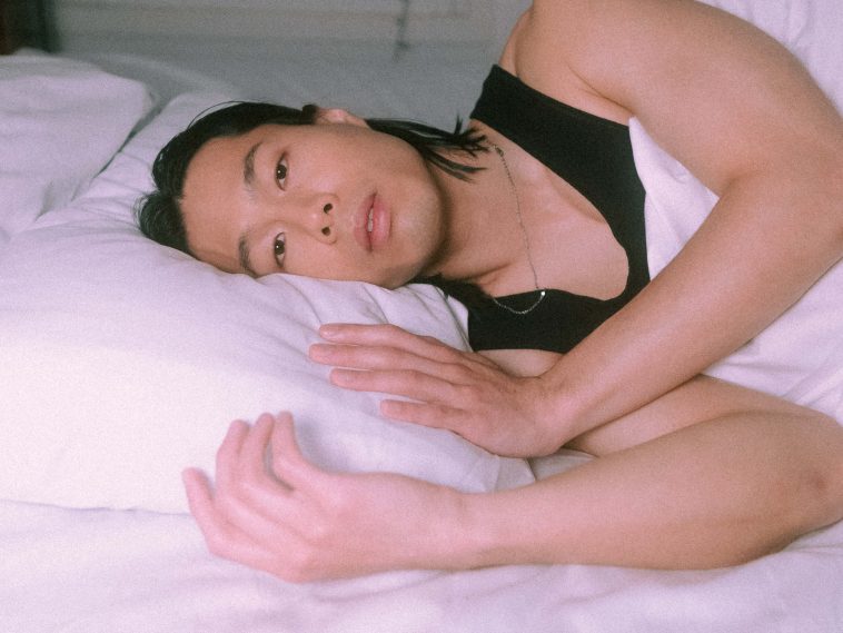 Promotional photo for "Pillow" which sees St. Humain lying on his side in a bed with his head on a pillow. His shoulder-length black hair is behind his ears and he's wearing a black vest. The bedding is white and the duvet is under his arm, while his hands are curled against the side of the pillow.