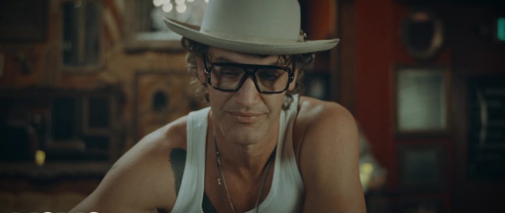 YouTube thumbnail of "The World's Gonna Love Me" which sees TJ Stafford wearing a white tank top, black square glasses, and a pale cream fedora, posing at the bar as he drinks his spirit.
