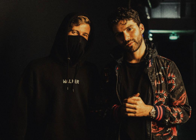 Promotional image for Alan Walker's remix of "All Around the World (La La La)" by R3HAB & A Touch of Class. Alan Walker and R3HAB are standing next to each other in a dark, dank warehouse, with Alan Walker wearing a black hoodie with the hood up and he is wearing a mask, so you can only see a bit of his hair, his forehead and his eyes. R3HAB is standing next to him, wearing an orange-black bomber jacket over a black t-shirt and he has his left arm out in front of him, He's looking at the camera and now has dark brown hair with a long stubble beard.