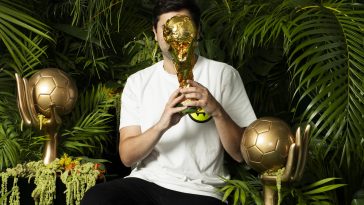 Promotional photo for "Globo" which sees BLVD. sitting on a throne amongst a jungle setting, holding the golden FIFA World Cup trophy in his hands and obscuring his face with it. He's wearing a white short-sleeve t-shirt and black jeans. There's two gold hand statues either side of him, holding a gold football.