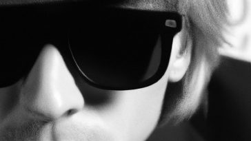 Promotional photo for "DEATH LETTER" which is a black and whited zoomed in headshot of Mortal Prophets frontman John Beckmann, where we see mainly just the left side of his face. He is wearing a hat - the brim of which we can just about see - with some sunglasses and his hair tucked behind his ear. The jacket of his suit can be seen on his shoulder.