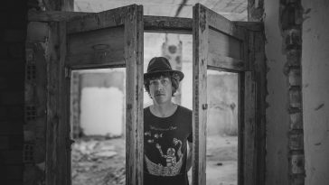 Promotional photo for "A Letter" which sees Gated Estates frontman Dan Linn-Pearl standing in a double-doorway with the doors half-open. He is standing there with a fedora hat and a t-shirt that has a night sky printed on it. The photo is in black and white.