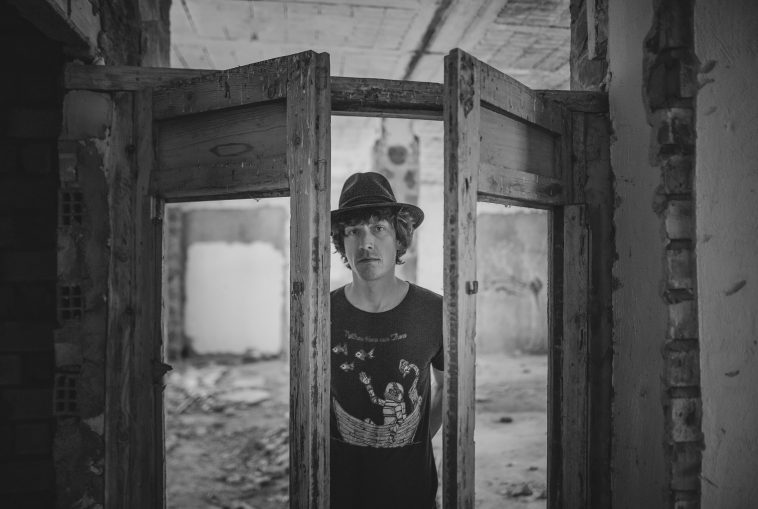 Promotional photo for "A Letter" which sees Gated Estates frontman Dan Linn-Pearl standing in a double-doorway with the doors half-open. He is standing there with a fedora hat and a t-shirt that has a night sky printed on it. The photo is in black and white.