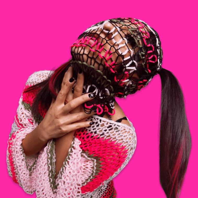 Promotional photo for "Pink Grenade" which sees LESS KILLJOY posing with a woolen multi-coloured facekini on with her long brown hair held up in a ponytail, and her head is tilted to top-left, allowing her ponytail to dangle down to the bottom right. She's wearing a cream woolen top that matches the facekini in inverted colours. There's a bright hot pink background emphasising LESS KILLJOY.