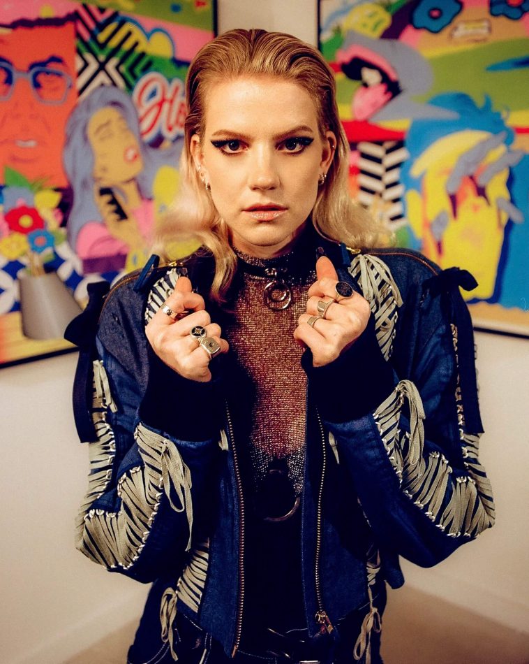 Promotional photo for "Battleground" which sees Levina in a blue bomber jacket pulling out towards the car with her hands gripped on the open zip. Her blonde hair is slicked back and it looks like she's posing in an art museum as there's art works ib a wall behind her.