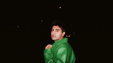 Promotional photo for "Caffeine High" which sees Sid Seth posing with the backdrop of the night sky. He is looking over one shoulder at the camera, wearing an oversized green plastic-like jacket.