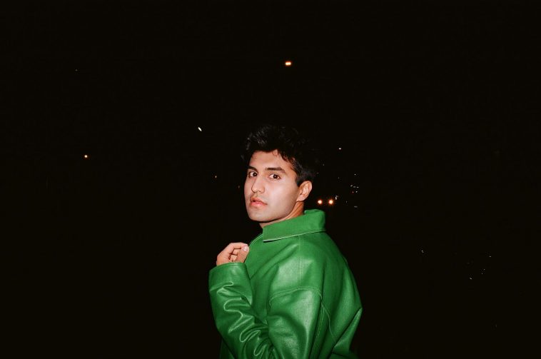 Promotional photo for "Caffeine High" which sees Sid Seth posing with the backdrop of the night sky. He is looking over one shoulder at the camera, wearing an oversized green plastic-like jacket.