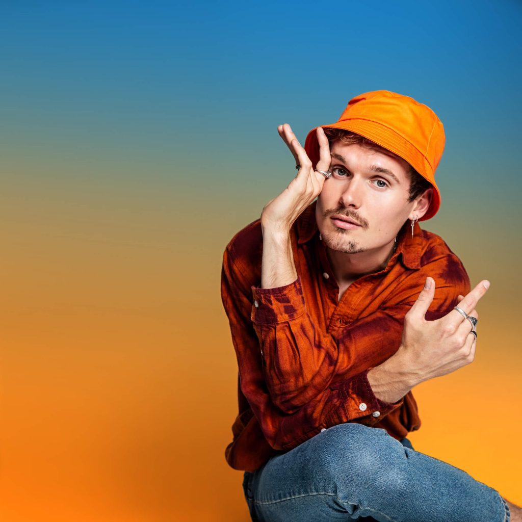 Promotional photo for "Heal" which sees Waiting for Smith also known as Harry Lloyd crossing his elbows in front of him, staring into the camera. He is wearing a light-orange bucket hat, a dark orange to black tie-dye shirt, and blue jeans. The background is an ombre colour starting from blue at the top to orange at the bottom.