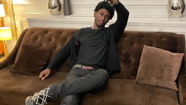 Freddie Wahter is sitting on a brown couch throwing the peach sign above his head with his fingers. He is wearing a black t-shirt, with a black hoodie, black jeans, and black trainers.