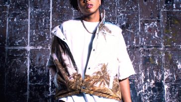Promotional photo for "Web of Lies" which sees La'Ron posing in front of a blue-tiled wall. He is wearing a white t-shirt underneath a white hoodie that has a beach and palm trees printed on the bottom in a sandy yellow, and blue cargo trousers with white trainers.