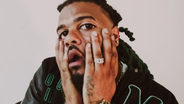 Promotional photo for "GANGOSO WORLD" which sees Moreno ITF looking shocked, with his hands on his cheeks, showing his silver bracelet and silver watch. He is wearing a black varsity jacket and his braided hair is behind him.