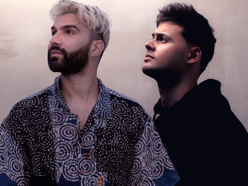 Promotional photo for "Sing Your Lullaby" which sees R3HAB on the left with his blonde hair and dark brown beard, wearing a swirl mosaic shirt that is in blue and dark blue vertical colouring with the swirls in white. He is looking to the top left of the image. Mike Williams is on the right, standing to the side, wearing a black hoodie and also looking upwards to the top left of the image.