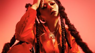 Promotional photo for "Unearthly World" which has a red filter over it and sees RayRay sitting on the floor with her right elbow touching her right knee, with her hand leant against her forehead. She's wearing a red jumpsuit and has her hair in bunches at the sides of her head, which trails down in braids.