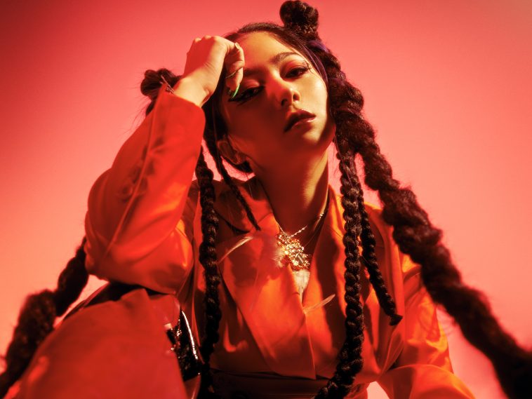 Promotional photo for "Unearthly World" which has a red filter over it and sees RayRay sitting on the floor with her right elbow touching her right knee, with her hand leant against her forehead. She's wearing a red jumpsuit and has her hair in bunches at the sides of her head, which trails down in braids.