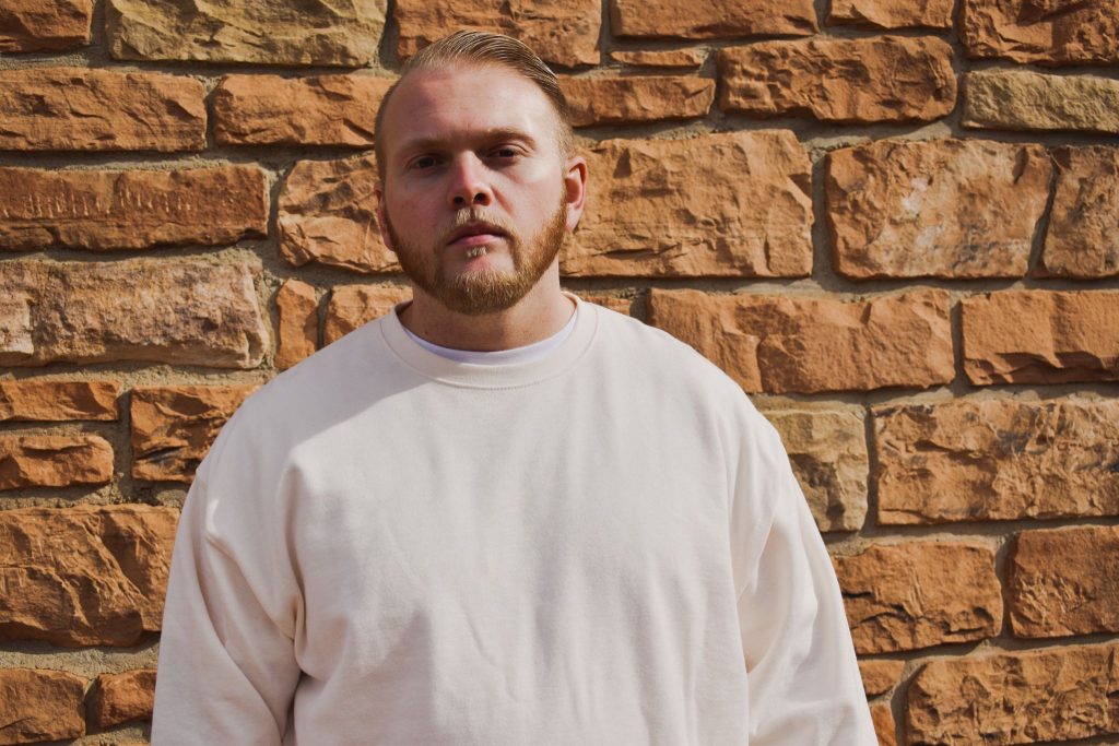 Promotional photo for "Meet Again" which sees Tay Toe standing against a brick wall wearing an off-white sweater. His hair is cut short and he has a trimmed beard.