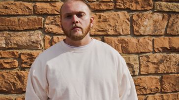 Promotional photo for "Meet Again" which sees Tay Toe standing against a brick wall wearing an off-white sweater. His hair is cut short and he has a trimmed beard.