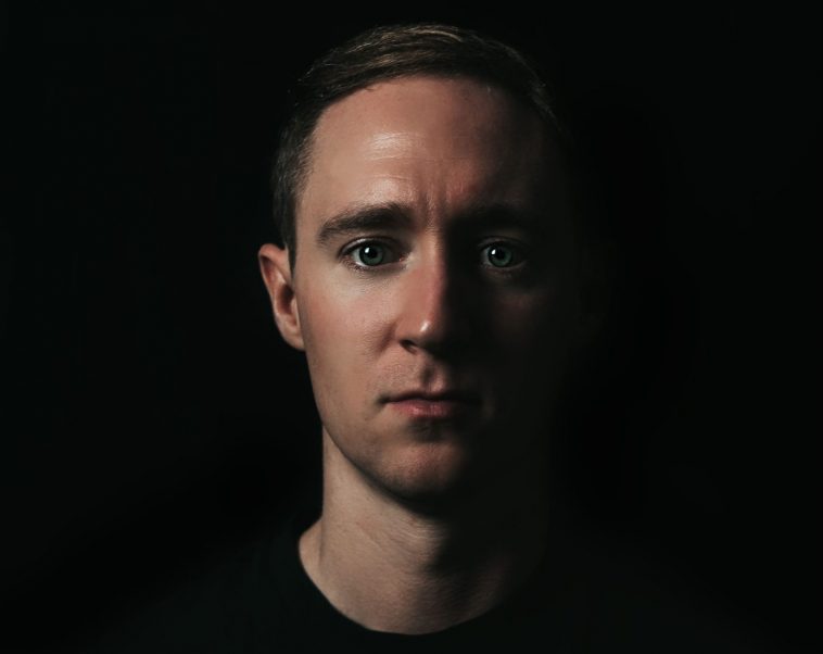 Promotional image for "I" which sees a headshot of Tontine against a black background. He is wearing a black t-shirt and the right side of his face is cast in shadow.