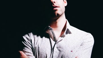 Promotional photo for "I Wish I Was Flawless, I'm Not" EP which sees BANNERS standing in front of a black backdrop, wearing a white long-sleeved shirt with the sleeves rolled up to his mid-lower arm. His arms are crossed and he has his head slightly tilted to the side.