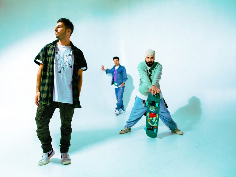 Promotional photo for "Anyway" featuring RuthAnne that shows the trio known as Cash Cash standing in various perspectives with one of them holding on to a skateboard. They're in a pale blue room, and one member is in the forefront wearing a white tee under an open green checkered shirt, cargo pants, and white Nike trainers. The one in the background is wearing a denim jacket over a white t-shirt that has a print on the front, paired with blue jeans and white trainers. The guy in the middle perspective is wearing a pale white beanie, a green jumper that matches the skateboard, blue sweatpants and dirty white trainers.