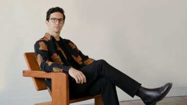 Promotional photo for "Slip Away" which sees Dan Croll sitting in a wooden relaxing chair with one leg crossed. He's wearing glasses and a black shirt that has patterned autumnal leaves on it, paired with black suit trousers and black smart shoes. He's in a grey room that is also fitted with grey carpet.