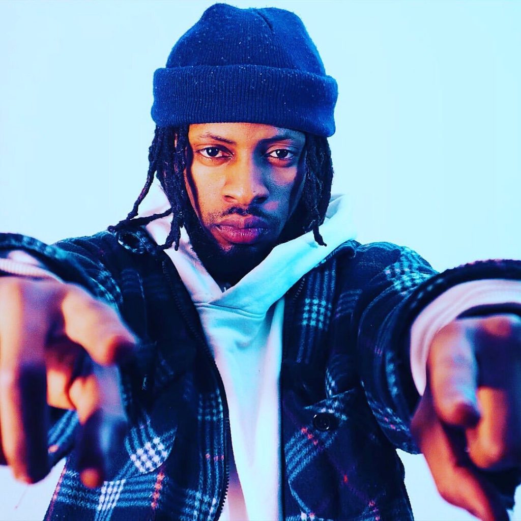 Promotional photo for "You Wasn't There" which sees 4REX with his arms out, pointing at the camera. He is wearing a blue beanie over his black dreads, a white hoodie under a blue and white checkered overshirt.