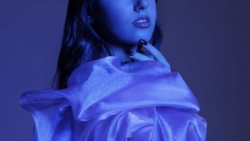 Promotional photo for "feeling blue" which sees a blue filtered image of Sindy Hoxha looking over her shoulder at the camera, with her long hair flowing down behind her. She is wearing a draped silk material which is bunched around the top-half of her body.