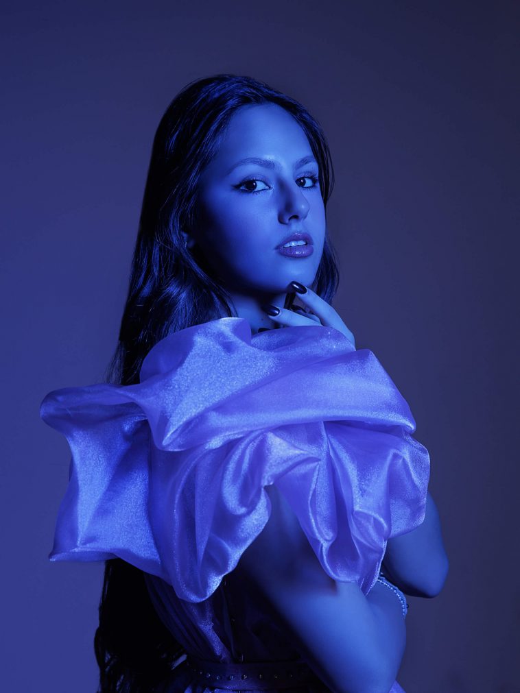 Promotional photo for "feeling blue" which sees a blue filtered image of Sindy Hoxha looking over her shoulder at the camera, with her long hair flowing down behind her. She is wearing a draped silk material which is bunched around the top-half of her body.