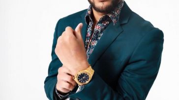 Promotional photo of Dylan Santana standing in front of a white background, wearing a teal green suit jacket over a blue floral shirt. He is adjusting his gold watch on hi left arm with his right hand, out in front of his chest.