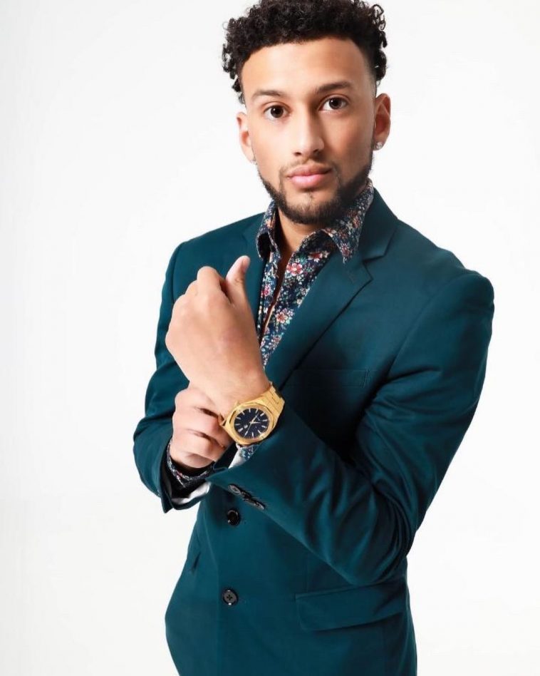 Promotional photo of Dylan Santana standing in front of a white background, wearing a teal green suit jacket over a blue floral shirt. He is adjusting his gold watch on hi left arm with his right hand, out in front of his chest.
