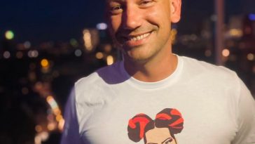 Promotional photo for Eurovision Song T-Shirts which sees Menno wearing an off-white t-shirt with an art image of Netta embossed on it, in an animation character style. He's on a balcony with a skyline behind him.