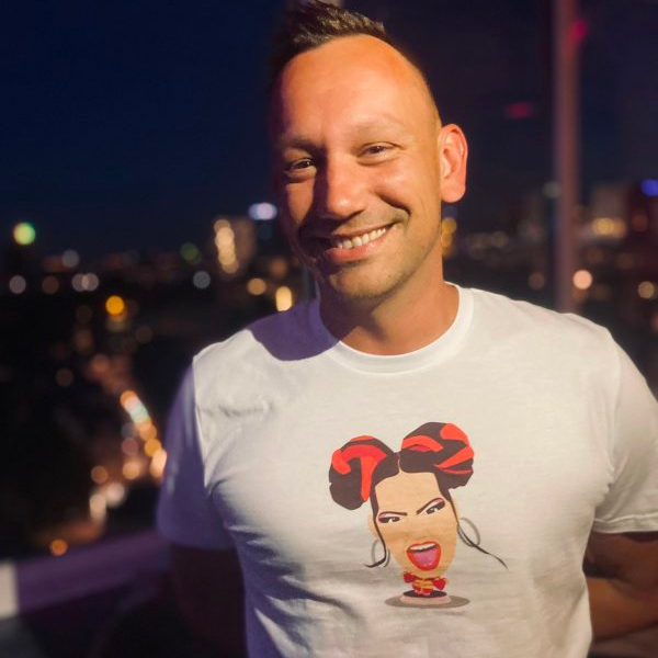 Promotional photo for Eurovision Song T-Shirts which sees Menno wearing an off-white t-shirt with an art image of Netta embossed on it, in an animation character style. He's on a balcony with a skyline behind him.
